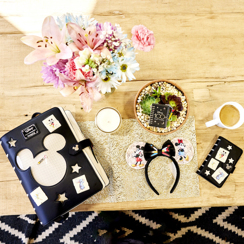 Wooden table with flowers and a cup of coffee with the Disney100 Sketchbook backpack, ear headband, pin, and wallet displayed on the table.